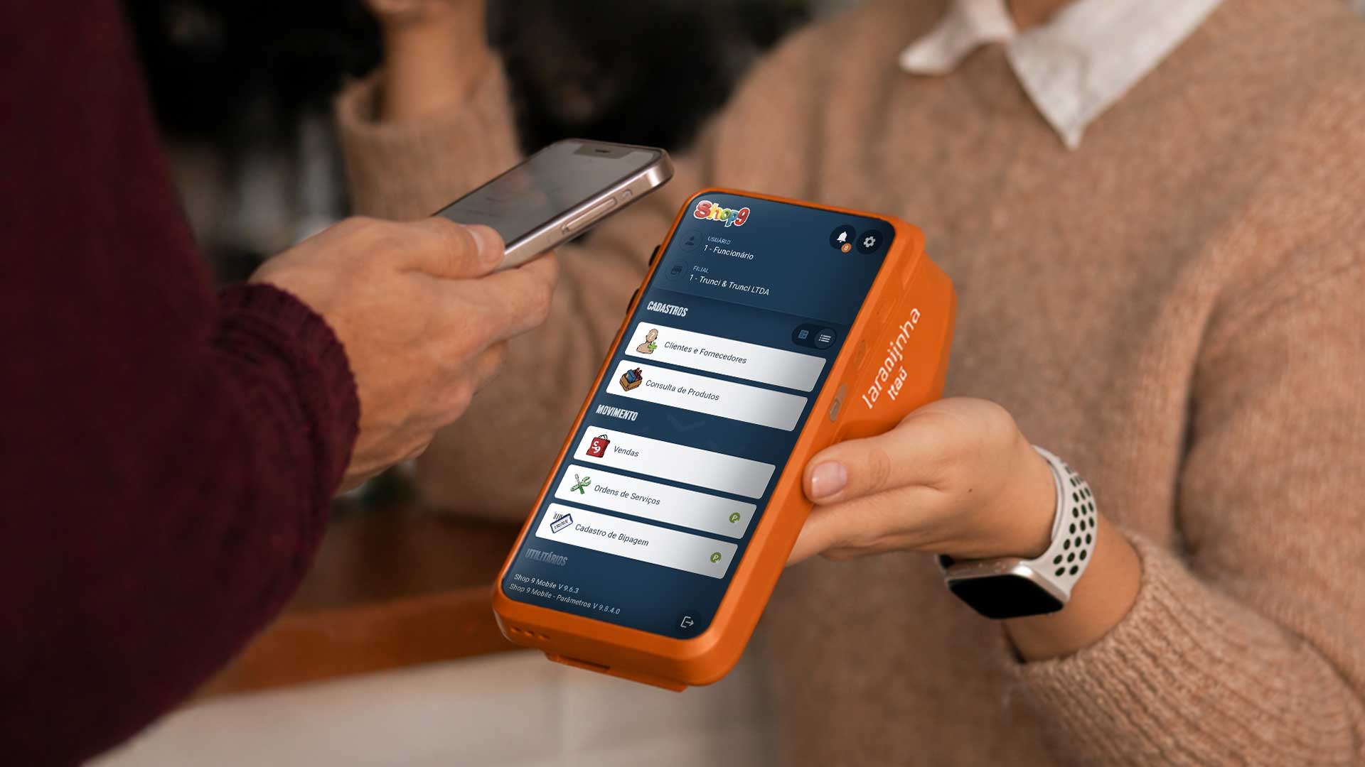 Ponto de vista do vendedor estendendo a maquina laranjinha com integração com o Shop 9 Mobile em direção ao cliente, que por sua vez estende o celular para efetuar pagamento do pacote que está em cima do balcão,